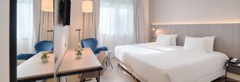 Bright hotel room at Hotel NH Roma Villa Carpegna with a double bed, white linens, desk, blue armchair, and window curtains
