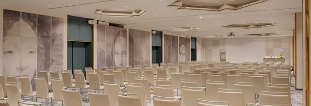 Conference room with rows of beige chairs, large wall murals of faces, and a white projector screen at Hotel NH Roma Villa Carpegna