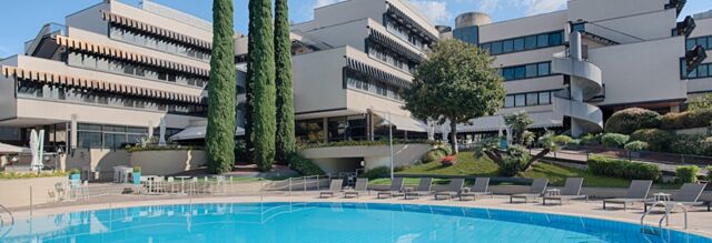 Outdoor pool area with lounge chairs, tables, trees, and modern white buildings at Hotel NH Roma Villa Carpegna