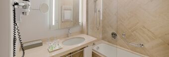 Bathroom with a sink, mirror, hairdryer, toiletries, bathtub with glass partition, and beige tiles at Hotel NH Roma Villa Carpegna