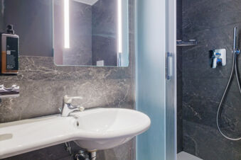 Bathroom sink with faucet, wall-mounted soap dispenser, illuminated mirror, and shower area with frosted glass door at B&B Hotel Ravenna