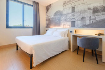 A clean white bed with four pillows beside a desk with a chair and a large window in B&B Hotel Ravenna room