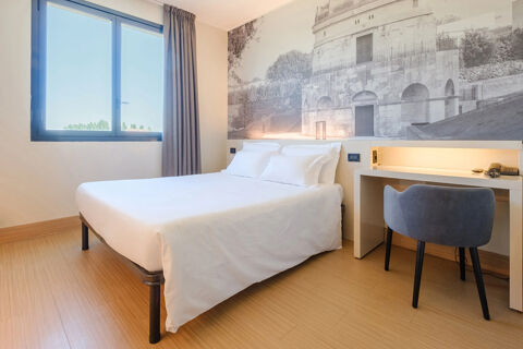 A clean white bed with four pillows beside a desk with a chair and a large window in B&B Hotel Ravenna room