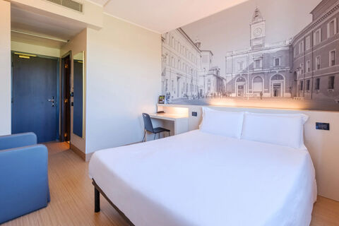 Modern hotel room at B&B Hotel Ravenna with a large bed, blue armchair, small desk with chair, and wall art mural