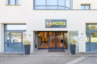 Entrance of B&B Hotel Ravenna with glass doors, two large gray planters with plants, and a yellow hotel sign above