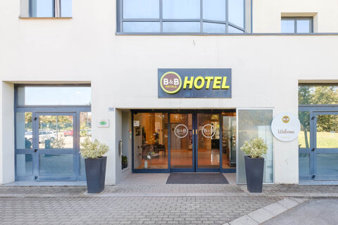Entrance of B&B Hotel Ravenna with glass doors, two large gray planters with plants, and a yellow hotel sign above
