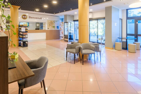 Lobby area of B&B Hotel Ravenna showing reception desk, snack stand, several grey chairs, tables, and large windows