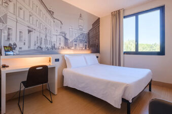 Room interior at B&B Hotel Ravenna featuring a double bed, a desk with chair, window with beige curtains, and wall mural
