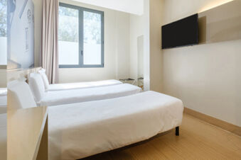 Two single beds with white linens are placed beside a window and a wall-mounted TV in a B&B Hotel Ravenna room