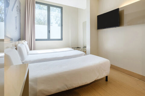 Two single beds with white linens are placed beside a window and a wall-mounted TV in a B&B Hotel Ravenna room