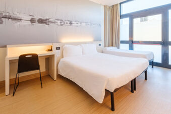 Hotel room at B&B Hotel Ravenna with two beds, a desk with a chair, a large wall mural of boats, and floor-to-ceiling windows