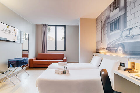 A modern hotel room at B&B Hotel Ravenna with two single beds, a brown sofa, a window, and a wall-mounted TV