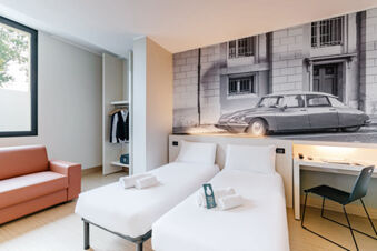 Modern hotel room at B&B Hotel Ravenna with two single beds, a brown couch, open closet, and a black and white car artwork on the wall