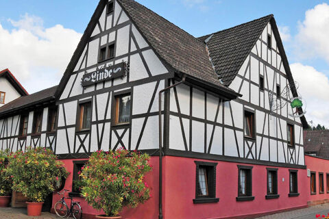 Hr&aacute;zden&yacute; dom s červen&yacute;m pr&iacute;zem&iacute;m a bielymi stenami hotela Gasthaus Zur Linde Diersburg, obklopen&yacute; kvetin&aacute;čmi a bicyklami.