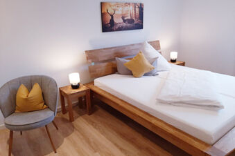 A wooden bed with white linens and multiple pillows next to bedside tables with lamps and a grey chair with yellow pillows at Hotel Gasthaus Zur Linde Diersburg