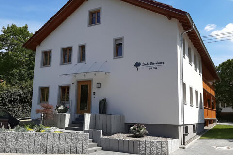 Fațada albă a hotelului Gasthaus Zur Linde Diersburg cu rame de ferestre maro și zona de intrare sub un cer &icirc;nsorit și plante.