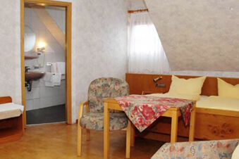 &Uacute;tuln&aacute; izba s dreven&yacute;m n&aacute;bytkom, kvetinov&yacute;mi text&iacute;liami a priľahlou k&uacute;peľňou v hoteli Gasthaus Zur Linde Diersburg so &scaron;ikmou strechou a osvetlen&yacute;m oknom.