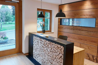 Reception desk with stacked wood design, black countertop, and two hanging lamps near a window at Hotel Gasthaus Zur Linde Diersburg