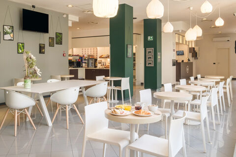 Dining area with white tables and chairs, food on a table, green columns, hanging lights, and a breakfast area at B&B Hotel Faenza