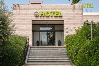 Entrance of B&B Hotel Faenza with glass doors, steps leading up, stone wall facade and green bushes on both sides