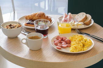 Breakfast on a round wooden table at B&B Hotel Faenza includes scrambled eggs with sausage, juice, coffee, cereal, croissant, and bread with cheese and ham