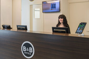 Reception desk with dark wood finish, a smiling female receptionist, two computer monitors, and a B&B Hotel Faenza logo on front