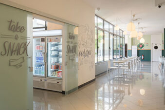 Modern hotel snack area with glass doors, a refrigerated display stocked with food and drinks, white tables and chairs, and pendant lights at B&B Hotel Faenza
