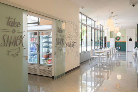 Modern hotel snack area with glass doors, a refrigerated display stocked with food and drinks, white tables and chairs, and pendant lights at B&B Hotel Faenza