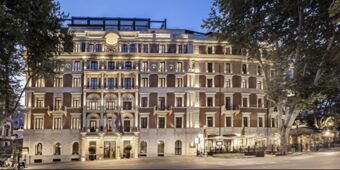 Lit fa&ccedil;ade of the InterContinental Rome Ambasciatori Palace building at dusk with detailed balconies and trees on both sides