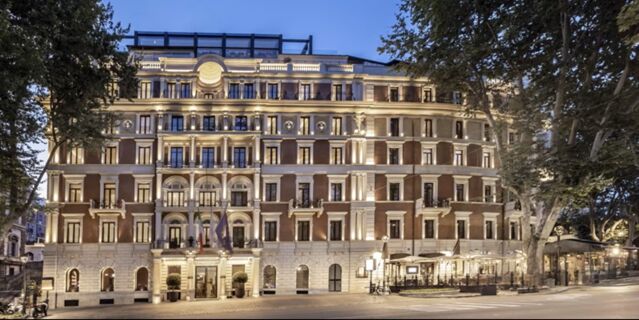 Lit fa&ccedil;ade of the InterContinental Rome Ambasciatori Palace building at dusk with detailed balconies and trees on both sides