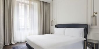 A bright hotel room at InterContinental Rome Ambasciatori Palace with a large bed, white linens, gray curtains, and hanging lights
