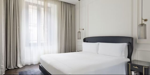 A bright hotel room at InterContinental Rome Ambasciatori Palace with a large bed, white linens, gray curtains, and hanging lights