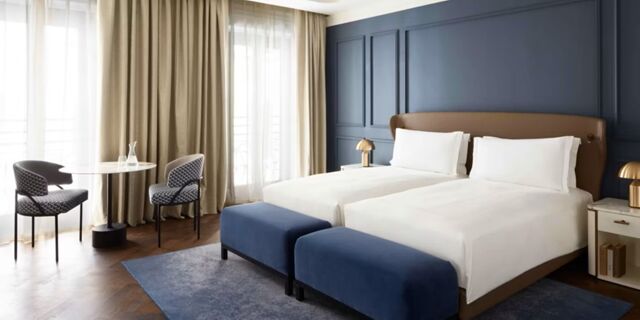 Two single beds with white linens, blue benches, a table with two chairs, and large curtains in InterContinental Rome Ambasciatori Palace bedroom