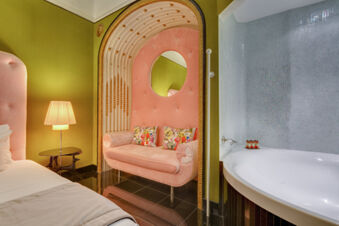 A pink tufted couch with floral pillows sits inside an ornate alcove next to a bed, lamp, and a white bathtub at Grand Hotel Palace