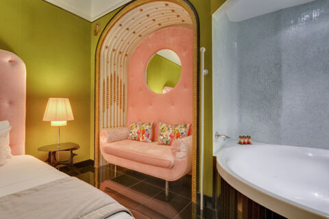 A pink tufted couch with floral pillows sits inside an ornate alcove next to a bed, lamp, and a white bathtub at Grand Hotel Palace