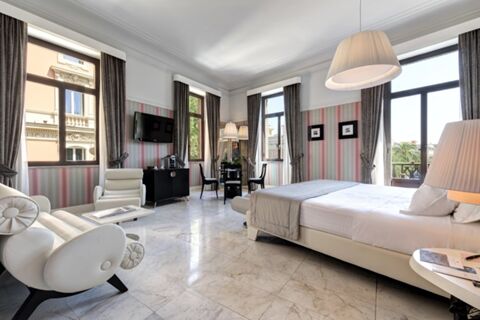 Bright hotel room at Grand Hotel Palace with large bed, two armchairs, marble floor, and multiple windows with curtains