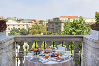 A balcony breakfast setup with juice, croissants, fruit, and coffee cups overlooking city buildings at Grand Hotel Palace