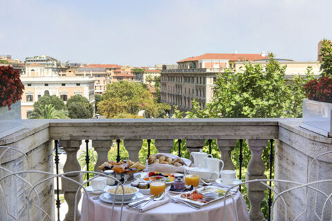 A balcony breakfast setup with juice, croissants, fruit, and coffee cups overlooking city buildings at Grand Hotel Palace