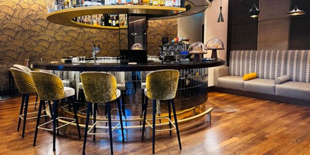 A curved black bar with bottles and coffee machines behind it, four velvet bar stools, decorative wall and a patterned sofa at Crowne Plaza Rome -St. Peter's