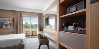 Bright hotel room at Crowne Plaza Rome -St. Peter's featuring a bed, desk with chair, balcony, and a wooden shelf with a safe and coffee setup