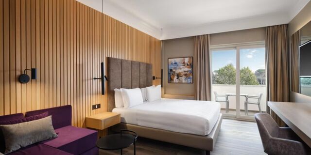 Modern hotel room at Crowne Plaza Rome -St. Peter's with a large bed, purple sofa, wooden panel wall, and balcony with chairs