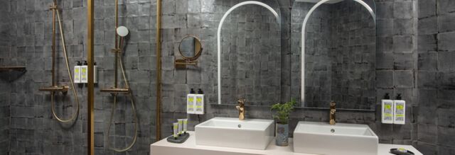 Modern bathroom with dual sinks, gold shower fixtures, grey tiled walls, mirrors, and toiletries at Hotel nhow Roma Vittorio Veneto