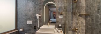 Modern bathroom with grey tiles, a white sink, gold fixtures, a mirror, hairdryer, and shower in Hotel nhow Roma Vittorio Veneto