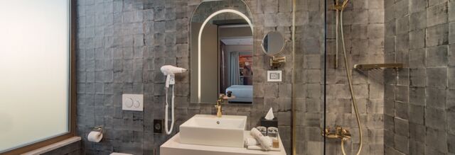 Modern bathroom with grey tiles, a white sink, gold fixtures, a mirror, hairdryer, and shower in Hotel nhow Roma Vittorio Veneto
