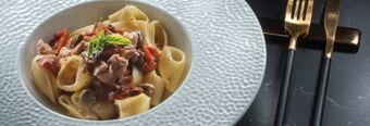 White textured bowl with pasta, pieces of meat, mushrooms, sun-dried tomatoes, and a basil leaf served at NH Collection Roma Centro with gold and black cutlery next to it