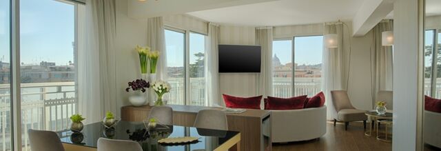 Spacious room at NH Collection Roma Centro with large windows, a dining table, chairs, flowers, and a curved sofa with red cushions