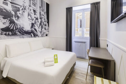 Double bed with white linens, towels, and a green sign, artwork of statues on wall, desk, chair, window, and curtains in B&B Hotel Roma Italia Viminale room