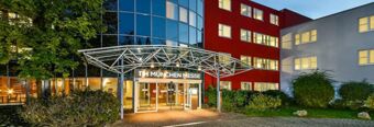 Entrance of NH Hotel M&uuml;nchen Neue Messe with glass doors under a metal canopy and adjacent red and white buildings at dusk