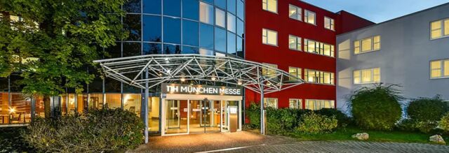 Entrance of NH Hotel M&uuml;nchen Neue Messe with glass doors under a metal canopy and adjacent red and white buildings at dusk