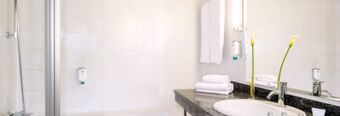 Bright bathroom with white tiles, a shower, neatly stacked towels, a round sink, and a flower at NH Hotel M&uuml;nchen Neue Messe